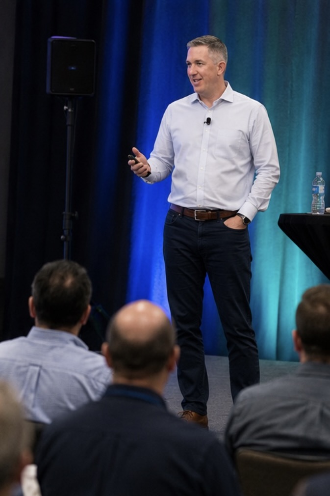Kevin Miller speaking to a seated audience, holding a presentation remote